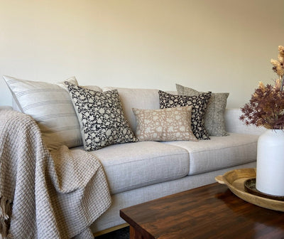 A light colored sofa with 5 decorator throw pillows, a lap blanket, coffee table with a white ceramic vase with dried floral arrangement on a wood tray. 