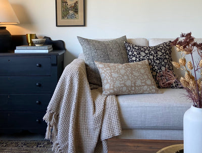 A light colored sofa with 5 decorator throw pillows, a lap blanket, coffee table with a white ceramic vase with dried floral arrangement on a wood tray. To the left of the sofa is a black chest of drawers with a lamp and stacked books.
