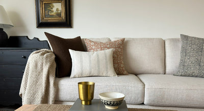 A sofa with 4 decorator throw pillows, a lap blanket across the left arm of the sofa. A large lumbar decorator throw pillow with an off white fabric and 6 vertical bars comprised of three lines are evenly spaced across the pillow. 