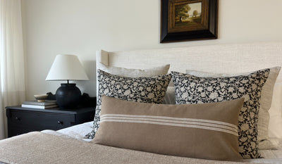 A bed with five decorator throw pillows. Two solid light gray pillow covers, two black and off white botanical print, one long lumbar in front with a solid brown fabric with a horizontal stripe comprised of 3 closely spaced lines.