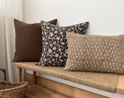 Three decorator throw pillows arranged on a simple wooden bench with the seat made with twine. One large brown pillow cover, a black and off white pillow cover, and a large lumbar pillow cover. 