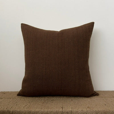 A single hand woven brown decorator throw pillow on a simple bench with a corded seat. 